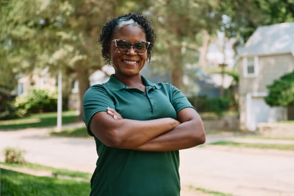 A confident Home Instead caregiver stands outdoors with arms crossed, smiling warmly, symbolizing professionalism and approachability.