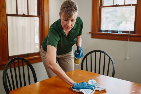 A Home Instead caregiver wearing gloves cleans a dining table with care, emphasizing thoroughness and attention to maintaining a clean, comfortable home.