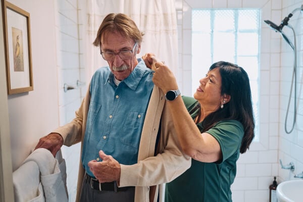 A Home Instead caregiver helps an elderly man adjust his sweater in a bathroom, emphasizing dignity and personalized care during daily routines.