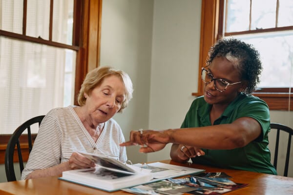 A Home Instead caregiver and an elderly woman review a photo album together at a dining table, sharing memories and stories.