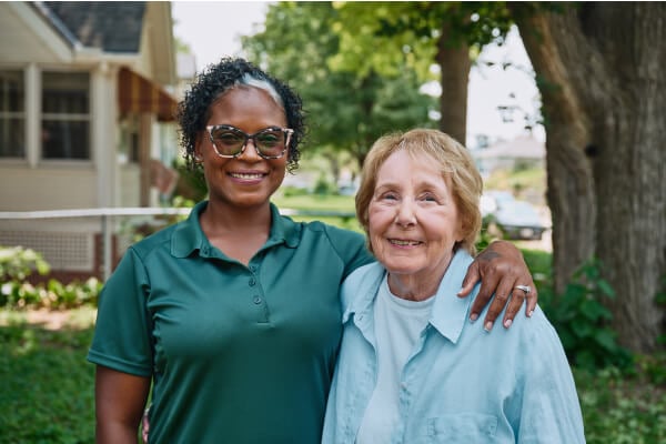 A Home Instead caregiver with an arm around an elderly woman outdoors, both smiling warmly, showcasing trust and companionship.