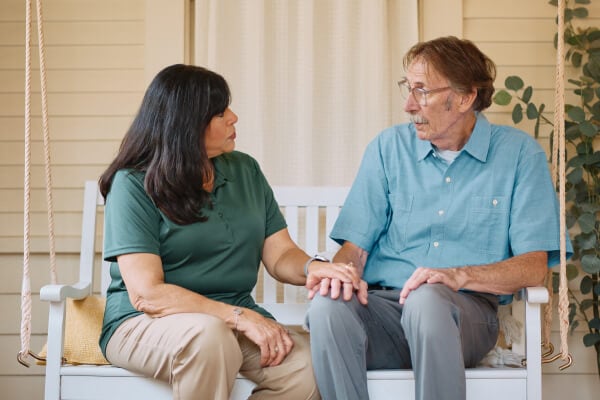 A Home Instead caregiver and an elderly man hold hands on a porch swing, fostering a sense of trust, reassurance, and emotional support.