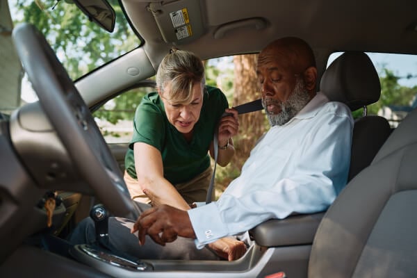 A Home Instead caregiver assists an elderly man in the passenger seat by fastening his seatbelt, ensuring safety and comfort during transportation.