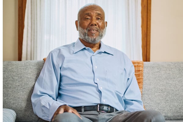 The same elderly man with a beard sitting on a comfortable sofa indoors, wearing a light blue shirt and appearing relaxed and at ease.