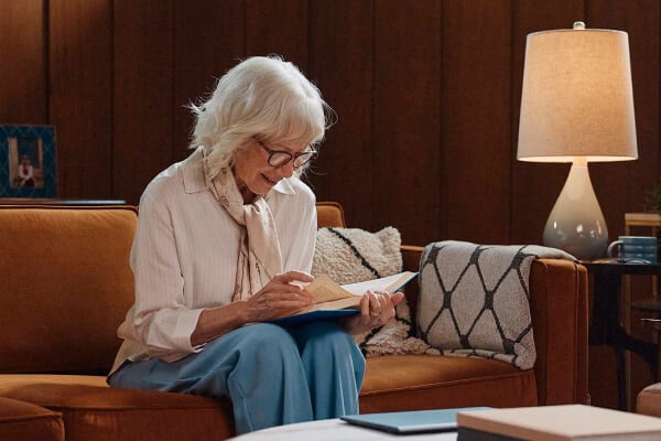 An older woman sits comfortably on a couch, reading a book under a warm lamp in a cozy living room, highlighting independence and peace of mind.