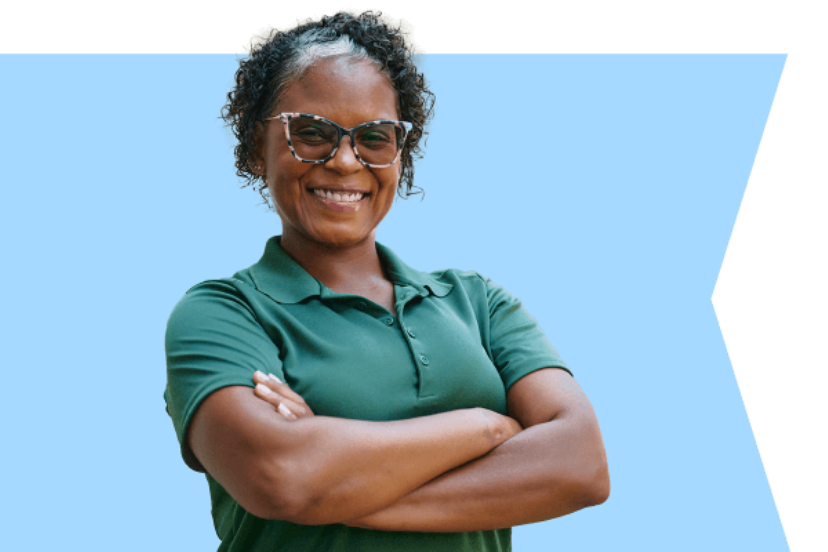 A Home Instead caregiver smiling in a green uniform with arms crossed, set against a blue flag-shaped background, symbolizing professionalism and trust.